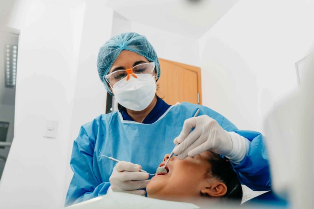 Dental professional in blue scrubs and protective gear performing an examination on a young patient in a clinical setting, emphasizing the importance of regular dental checkups.