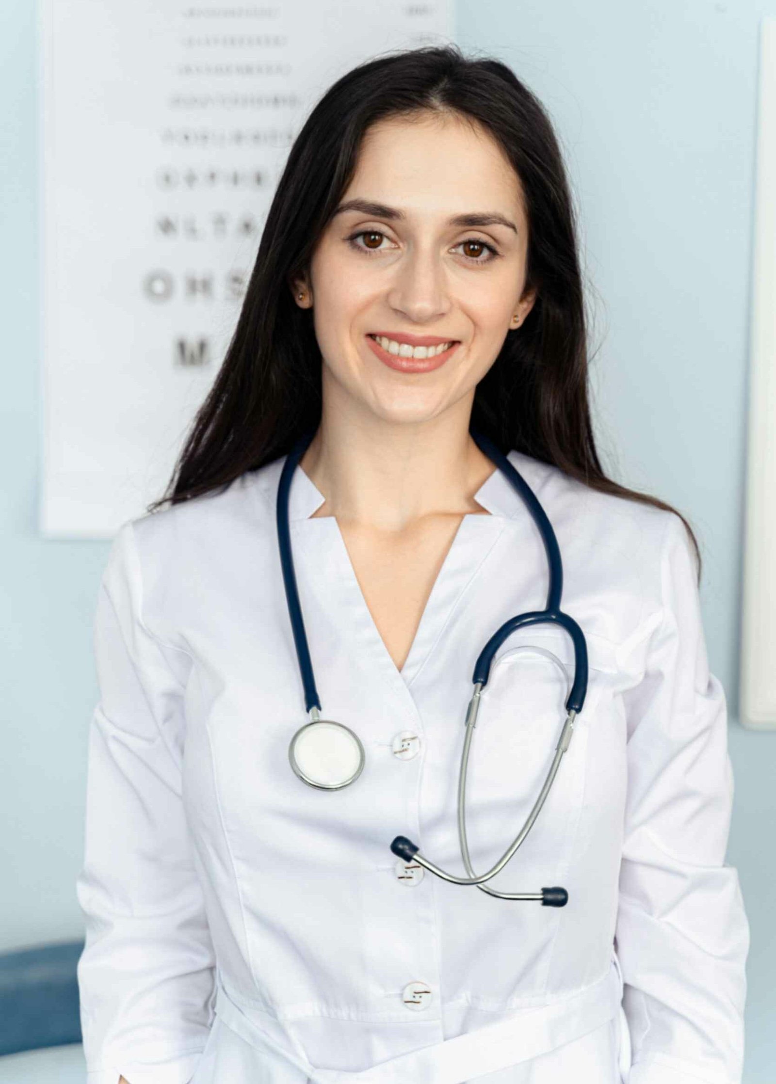 Smiling female dentist wearing a white coat and stethoscope, standing in a dental office with an eye chart in the background, representing Peak Performance Health's team of qualified professionals.