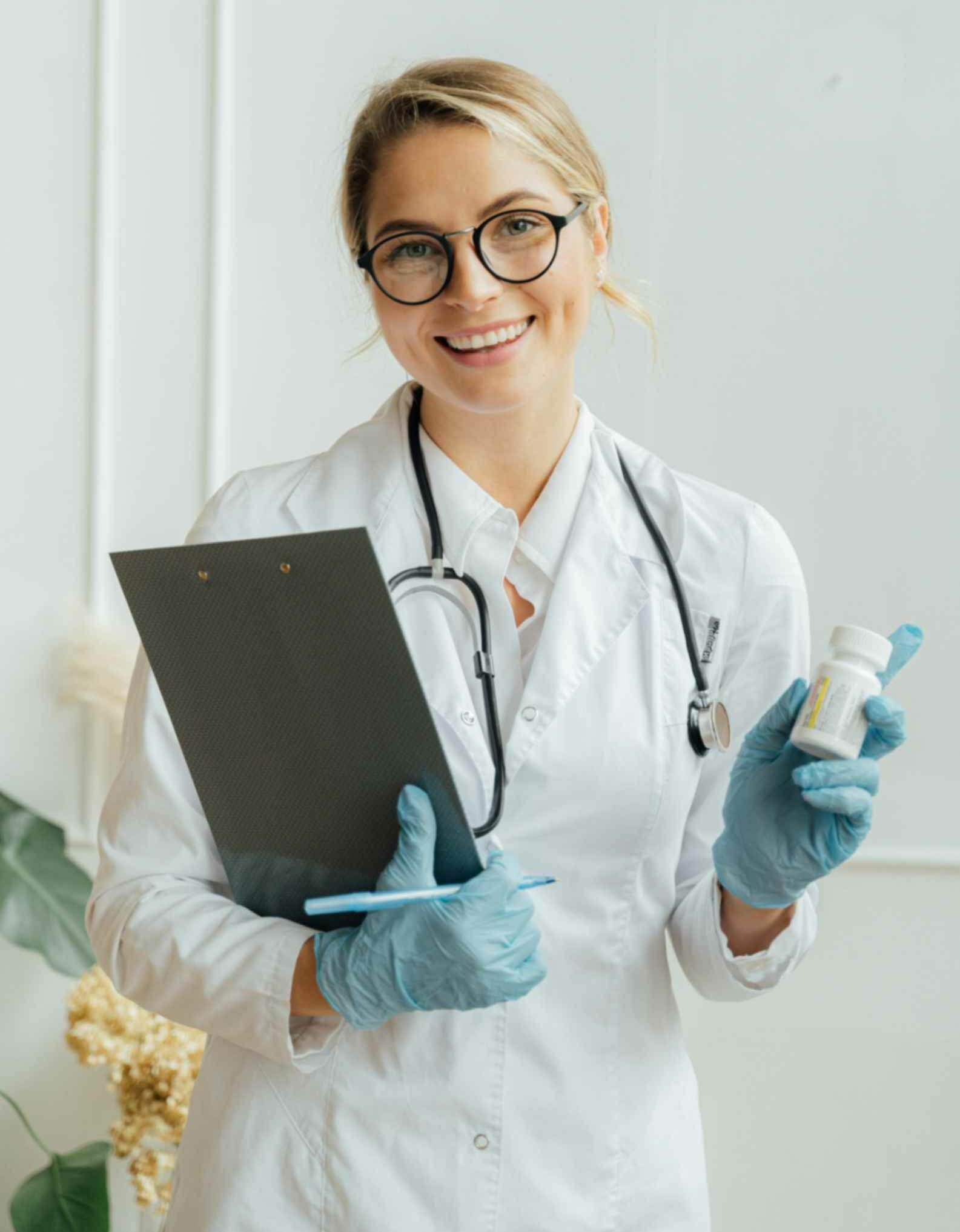 Healthcare professional in a white coat holding a clipboard and prescription bottle, smiling, representing qualified doctors and patient care at Peak Performance Health and Wellness.